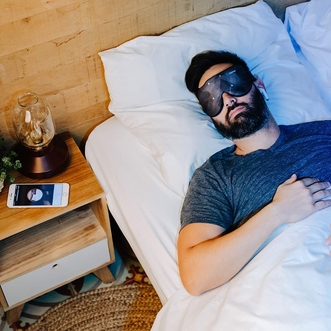 Homme Au Lit Avec Masque connecté HYPNOS de Dreaminzzz Noir A Côté d'Une Lampe et De Son Smartphone